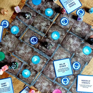 Op de afbeelding: Een bordspel met bruine tegels, blauwe en witte fiches, dobbelstenen en kaarten. De kaarten bevatten tekst zoals "Burning Fingers" en "Veil of Silence". De spelstukken bevatten schedel- en vleugelsymbolen.