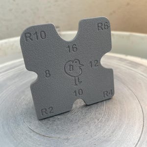 Può includere: Uno strumento da ceramica quadrato grigio con angoli arrotondati e sezioni ritagliate. Lo strumento ha i numeri 2, 4, 6, 8, 10, 12 e 16, insieme alle lettere R4, R6 e R10. Un piccolo logo di uccello è al centro.