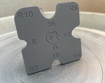 May include: A gray, square pottery tool with rounded corners and cut-out sections. The tool has the numbers 2, 4, 6, 8, 10, 12, and 16, along with the letters R4, R6, and R10. A small bird logo is in the center.
