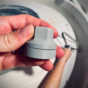 Könnte beinhalten: Ein hellgraues, zylindrisches Töpferwerkzeug mit facettierter Oberseite und gerillter Basis. Das Werkzeug wird in einer Hand gehalten, mit einem Rippenwerkzeug mit Holzgriff und einer Töpferscheibe im Hintergrund. Das Werkzeug scheint zum Formen von Ton zu dienen.