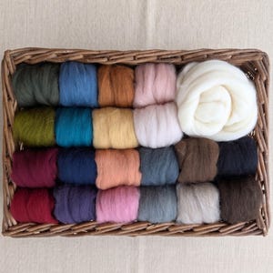 Op de afbeelding: Een rechthoekige gevlochten rieten mand met tweeëntwintig individuele bundels wolvilt in verschillende kleuren, waaronder tinten groen, blauw, bruin, roze, rood, grijs en een grote, opgerolde bundel wit wolvilt.
