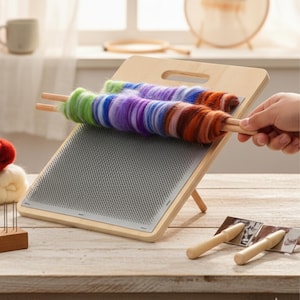 May include: A wooden felting board with a grey needle surface and a wooden stand. Two colourful yarn rolls are on the board, with a hand holding one of the wooden dowels. Additional wooden tools and yarn are on the wooden table.