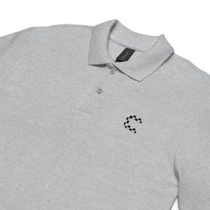 Puede incluir: Una camisa polo de manga larga gris con una letra "C" a cuadros bordada en el pecho derecho. La camisa tiene un cuello abotonado y una tapeta de tres botones.