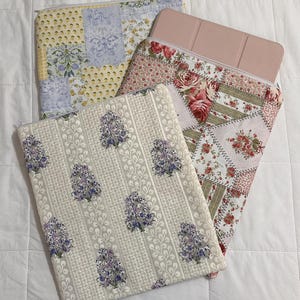 Könnte beinhalten: Drei gesteppte Stoffhüllen mit Blumen- und Patchworkmustern. Eine Hülle hat einen cremefarbenen Hintergrund mit lila Blumenmustern. Eine andere hat ein gelbes und blaues Patchwork-Design. Eine dritte Hülle hat ein rosa Blumenmuster.
