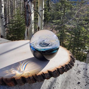 May include: A glass orb with a swirling blue and black design sits on a wooden slice. The orb is clear and has a textured surface. The background is a blurred image of trees.