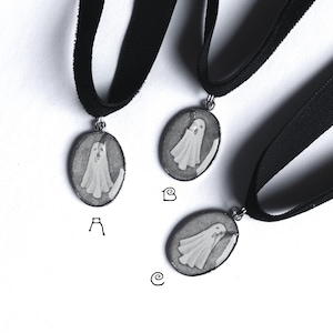 May include: Three black velvet chokers with oval pendants featuring a white ghost design. The ghost is set against a grey background. The pendants are attached to the choker with a silver-coloured bail.