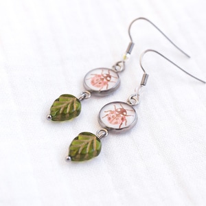 May include: Dangle earrings with a ladybug design. Each earring features a small, round charm with a ladybug illustration, connected to a green leaf-shaped bead. The earrings have silver-coloured ear wires.