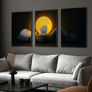 May include: Three framed art prints featuring a cartoon figure in a dark setting. The central print shows the figure with a gloved hand in front of a large yellow circle. The other prints show the figure in different poses. The prints are displayed above a gray sofa.