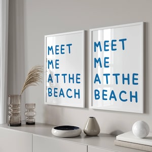 Könnte beinhalten: Zwei weiße gerahmte Drucke mit dem Text "Meet me at the beach" in blauer Schrift. Die Drucke hängen an einer weißen Wand über einer grauen Kommode.