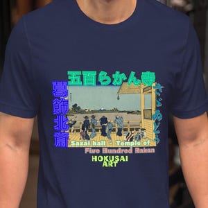 Pode incluir: Uma camiseta azul marinho com uma estampa gráfica de uma impressão japonesa em madeira que retrata um templo com pessoas em pé em uma varanda. O texto "Five Hundred Rakan" e "Hokusai Art" está impresso abaixo da imagem.