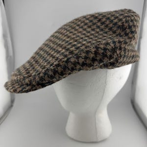 May include: A flat cap featuring a brown and black houndstooth pattern. The cap is constructed from a textured, wool-like fabric with a rounded crown and a short, stiff brim extending from the front, displayed on a plain white mannequin head.