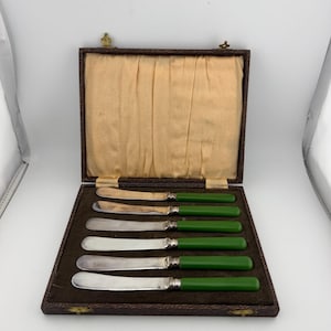May include: A set of six butter knives with smooth, solid green handles and silver-toned metal blades. The knives are arranged horizontally inside a rectangular, dark brown hinged presentation case with a cream-colored fabric interior lining.