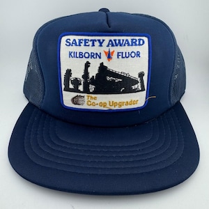 May include: A navy blue trucker hat with a mesh back and a white rectangular patch on the front. The patch features the text SAFETY AWARD KILBORN FLUOR and The Co-op Upgrader, along with a black silhouette graphic of an industrial facility.