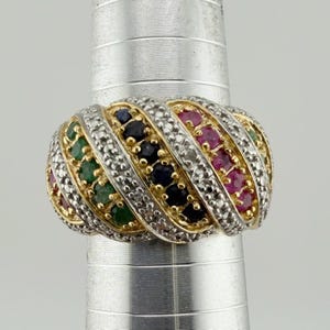May include: A multi-row gold ring featuring diagonal channels of round-cut green emeralds, dark blue sapphires, and bright red rubies, separated by rows of small white diamonds set in a textured, metallic band structure.