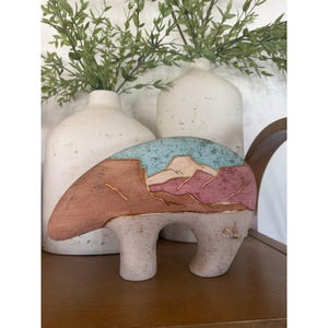 May include: A ceramic bear figurine with a landscape design. The bear has a light pink body and a turquoise and pink landscape design on its back. Gold accents outline the landscape. The figurine is on a wooden surface.
