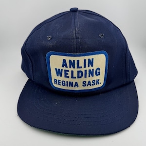 May include: A navy blue baseball cap with a flat brim. The cap features a rectangular patch with the text "ANLIN WELDING REGINA SASK." in white letters on a cream-colored background. The cap has a rounded crown and a slightly worn appearance.