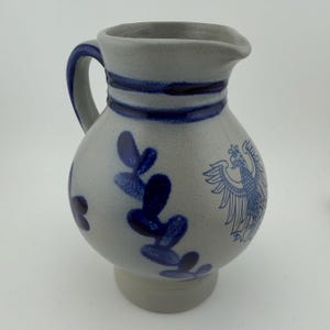 May include: A gray stoneware pitcher with a rounded body and a single handle. The exterior features cobalt blue hand-painted designs, including a detailed eagle emblem on the front and a leafy vine pattern, with two horizontal blue bands at the neck.