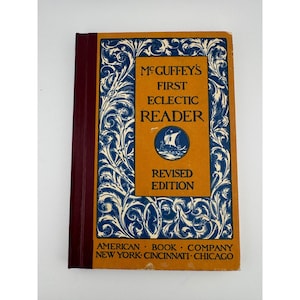 Może przedstawiać: Zabytkowa książka zatytułowana "McGuffey's First Eclectic Reader, Revised Edition". Okładka jest pomarańczowa z niebiesko-białymi dekoracyjnymi wzorami. Grzbiet jest bordowy. Wydane przez American Book Company.