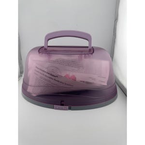 May include: A purple plastic cake carrier with a handle on top. The carrier has a clear, domed lid and a gray base. Inside the carrier are papers with text. The carrier is designed to transport and store baked goods.