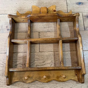 Puede incluir: Un estante de pared de madera teñida en color marrón medio con un remate decorativo en forma de lazo, seis compartimentos rectangulares dispuestos en dos filas y un borde inferior festoneado con tres pequeños ganchos de madera.