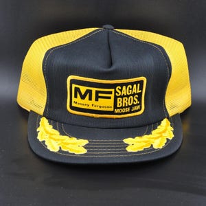 May include: Black and yellow trucker hat with the text "MF SAGAL BROS. MOOSE JAW" on the front. The hat has a mesh back and a curved brim with yellow leaf accents. The hat is a vintage style.