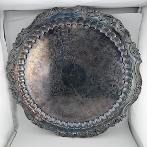 May include: A round silver-toned serving tray featuring an intricate engraved floral pattern across the center. The tray has a scalloped inner rim and a wide, decorative border with embossed scrollwork and leaf motifs along the outer edge.