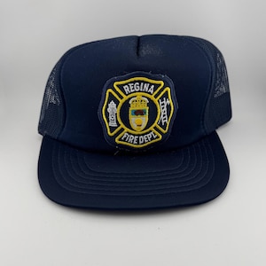 May include: Navy blue trucker hat with a mesh back and a curved brim. The front features a patch with the words "Regina Fire Dept" surrounding a crest with a crown, fire hydrant, and axe.
