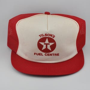 Könnte beinhalten: Rote und weiße Trucker-Mütze mit dem Text "TILSON'S FUEL CENTRE". Die Mütze hat einen roten Schirm und eine Netzrückseite, mit einer weißen Frontplatte mit dem Logo. Eine klassische Mütze im Vintage-Stil.