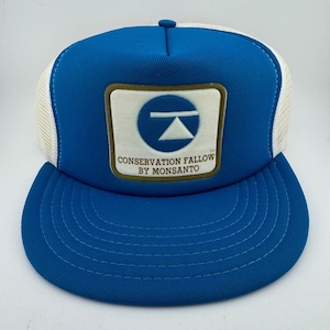 May include: A bright blue trucker hat with white mesh side panels and a structured front panel. A square patch on the front features a blue circle with a white triangle and horizontal bar, with the text CONSERVATION FALLOW BY MONSANTO.