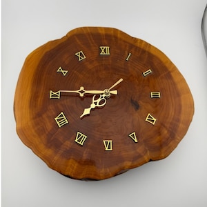 May include: A circular wooden clock with a natural edge. The clock face is made from a cross-section of a tree trunk, showing the wood grain. Gold Roman numerals mark the hours, and the clock hands are also gold. The clock has a rustic, natural aesthetic.