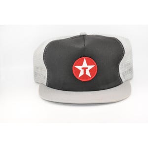 May include: A gray and black trucker hat with a red and white star logo on the front. The hat has a mesh back and a flat brim. The logo is a red circle with a white star and the letter "T" inside.