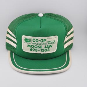 May include: Green and white trucker hat with a mesh back and a curved brim. The front panel features a white rectangular patch with the text "CO-OP IMPLEMENTS MOOSE JAW 693-1303" and the "DEUTZ ALLIS" logo. The hat has white stripes on the sides.