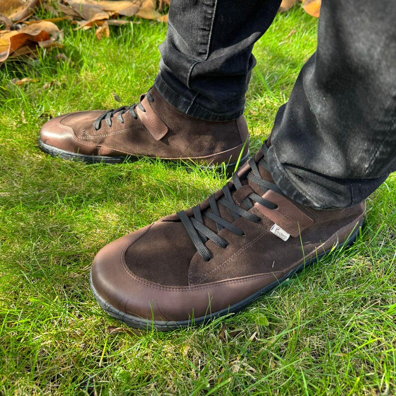 May include: Brown suede high-top sneakers with black laces and a brown leather sole. The shoes are worn by a person standing on grass.