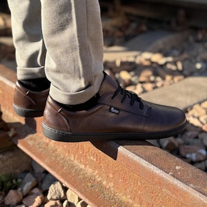 Puede incluir: Zapatos marrones de cuero con cordones y suela de goma negra. Los zapatos son usados por una persona de pie sobre una vía de tren oxidada.