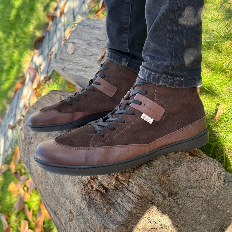 May include: Brown suede high-top sneakers with black laces and a brown leather toe. The shoes have a black rubber sole and a brown leather strap across the top.