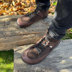 May include: Brown leather high-top boots with black laces. The boots are made of suede and leather and have a round toe. The boots are worn by a person standing on a wooden log.