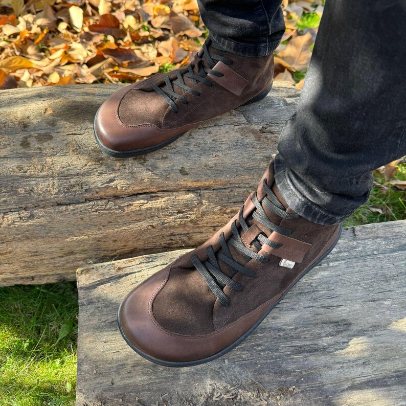 May include: Brown leather high-top boots with black laces. The boots are made of suede and leather and have a round toe. The boots are worn by a person standing on a wooden log.