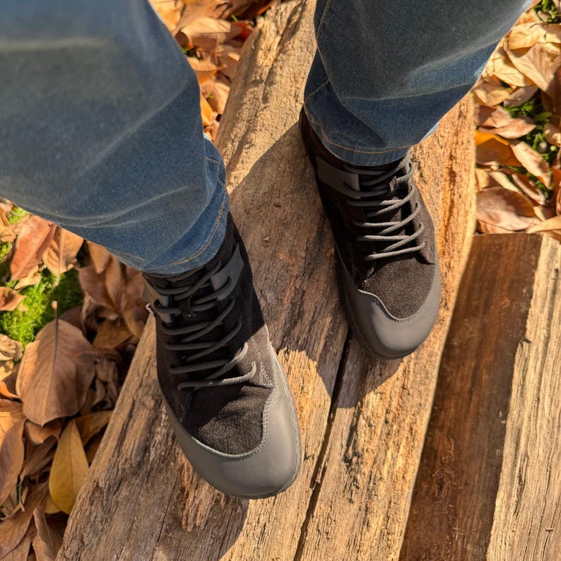 Pu&ograve; includere: Un paio di stivali neri in pelle scamosciata con lacci neri. Gli stivali sono indossati da una persona che indossa jeans blu e sono su una superficie di legno.
