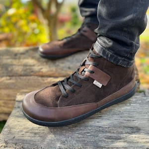 May include: Brown suede high-top sneakers with black laces and a brown leather toe cap. The shoes have a black rubber sole and a brown leather strap with the word "Gaimo" on it.