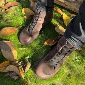 May include: A pair of brown leather ankle boots with black laces. The boots are worn by a person standing on a bed of green grass and fallen leaves.