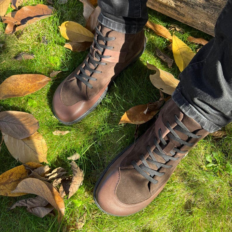 May include: A pair of brown leather ankle boots with black laces. The boots are worn by a person standing on a bed of green grass and fallen leaves.
