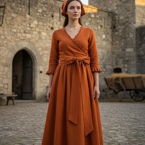 Op de afbeelding: Een oranjebruine wikkeljurk met bijpassende hoofdbedekking. De jurk heeft een V-hals, lange mouwen met ruches en een grote strik in de taille. Het model staat voor een stenen gebouw.