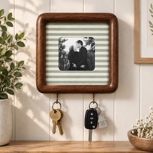 May include: A square dark wood wall-mounted key holder featuring a central photo frame with a black and white image of two people. The frame background has horizontal sage green and white stripes. Two metal hooks hold sets of keys below.