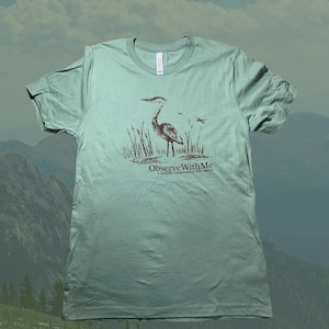 Könnte beinhalten: Hellgrünes T-Shirt mit einer braunen Illustration eines Reihers im Wasser und Schilf. Der Text "ObserveWithMe a wildlife conservation film series" ist darunter aufgedruckt. Das T-Shirt hat einen Rundhalsausschnitt und kurze Ärmel.