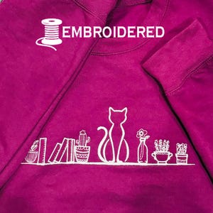 Puede incluir: Una sudadera magenta con un diseño bordado en blanco. El diseño incluye una silueta de gato, plantas en macetas y libros. La palabra "EMBROIDERED" está encima del diseño, y una bobina de hilo está en la esquina superior izquierda.
