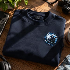 Peut inclure: Un sweat-shirt bleu marine à col rond avec un motif circulaire brodé sur la poitrine gauche. Le design représente deux poissons koï, l'un blanc et l'autre noir, nageant dans un tourbillon de vagues d'eau bleues et blanches.