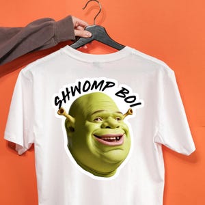 Peut inclure: T-shirt blanc avec un grand graphisme du visage de Shrek et le texte "SHWOMP BOI" en noir. Le graphisme vert de Shrek sourit. Le t-shirt est suspendu à un cintre noir sur un fond orange.