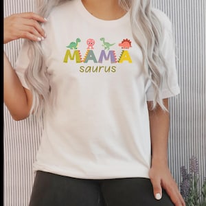 Könnte beinhalten: Ein weißes T-Shirt mit kurzen Ärmeln und einem Grafikdruck mit dem Text MAMA saurus in bunten Blockbuchstaben. Das Design zeigt vier kleine Cartoon-Dinosaurier in Grün, Pink und Orange, die über dem Wort MAMA platziert sind.