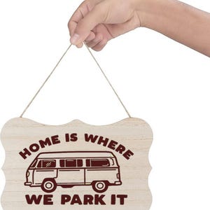 May include: Wooden sign with a light wood grain finish, featuring a brown illustration of a camper van and the text "HOME IS WHERE WE PARK IT." The sign is held by a natural twine string.