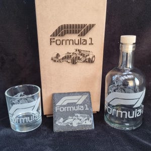 Könnte beinhalten: Ein Geschenkset zum Thema Formel 1 mit einer transparenten Glasflasche, einem Becher und einem Schieferuntersetzer. Das Set enthält eine Flasche mit Korken und eine Box mit dem Formel 1-Logo und einem Auto.
